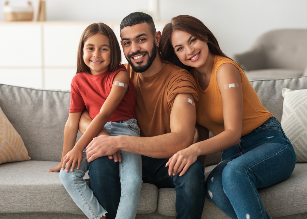 Coronavirus Vaccination Advert. Happy Vaccinated Family Of Three People Showing Arm With Sticking Plaster After Covid-19 Vaccine Vax Injection Posing Sitting On Couch In Living Room, Smiling To Camera