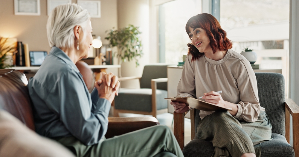 Psychologist, chat and senior woman with notes and smile from mental health assessment in consultation. Writing, support and care with counseling, psychology and evaluation with advice in office