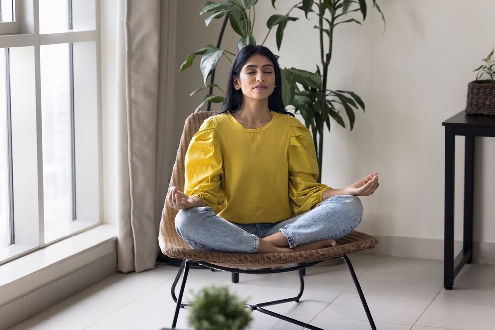 Calm Indian woman meditating at home seated in cozy armchair, practice encourages relaxation, enhance emotional well-being, promotes feelings of emotional stability, relieving stress, reduce anxiety