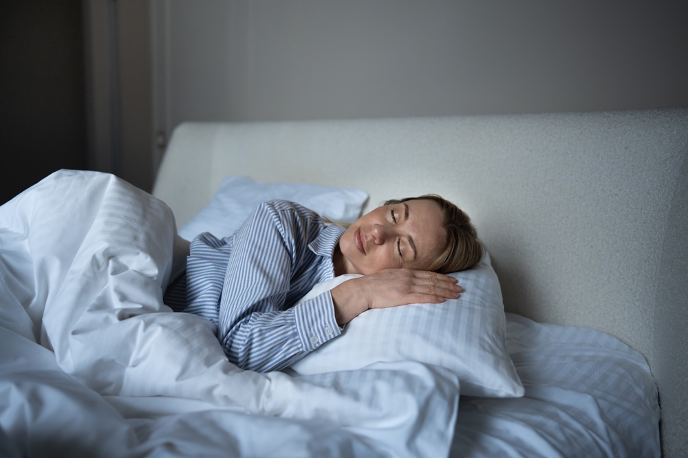 Calm middle aged woman sleeping in soft striped pajamas, snuggled into white blanket. Serene mature female resting in bedroom feeling harmony, emotional comfort. Self care, sleep hygiene, morning vibe
