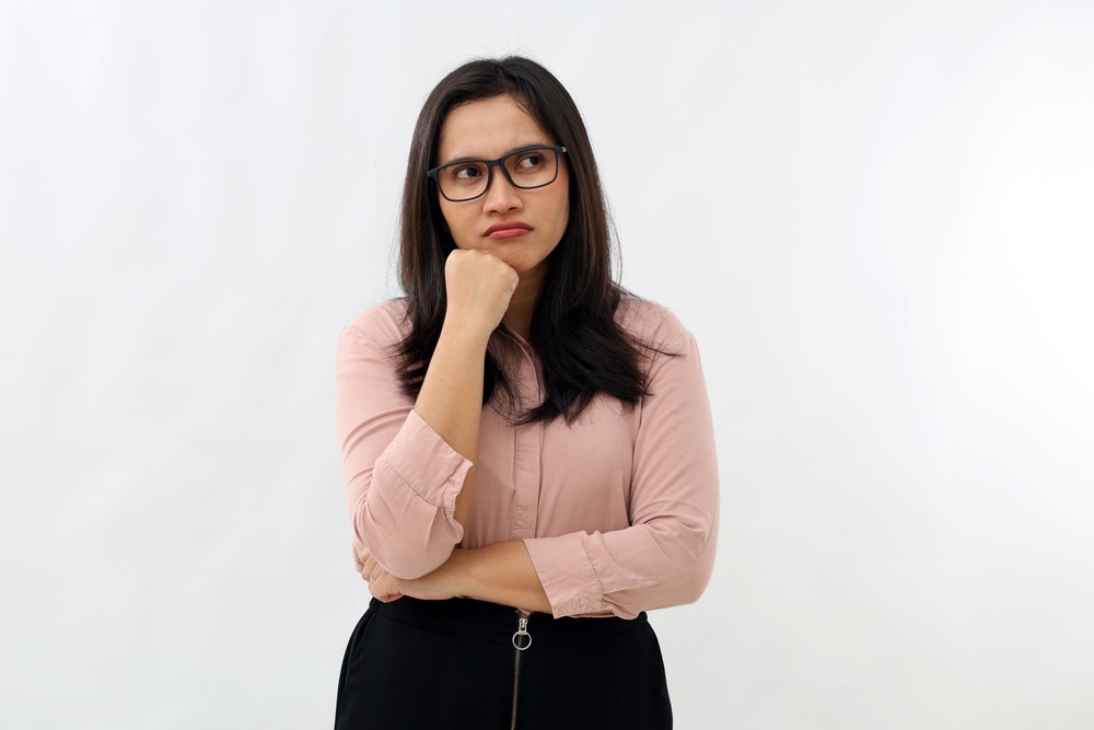 Young Asian woman standing with folded arms and supporting chin with hand, annoyed, angry, thinking expression, wearing formal shirt, glancing sideways into empty space, isolated on white background.
