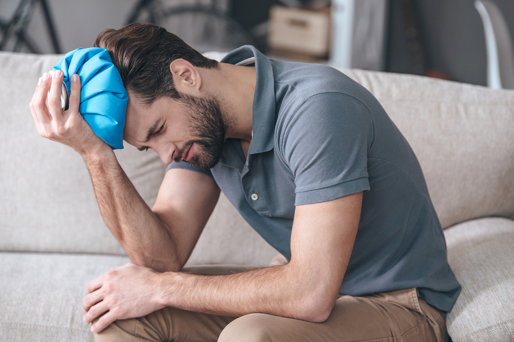 Feeling terrible headache. Frustrated young man holding ice bag on his head while sitting on the couch at home