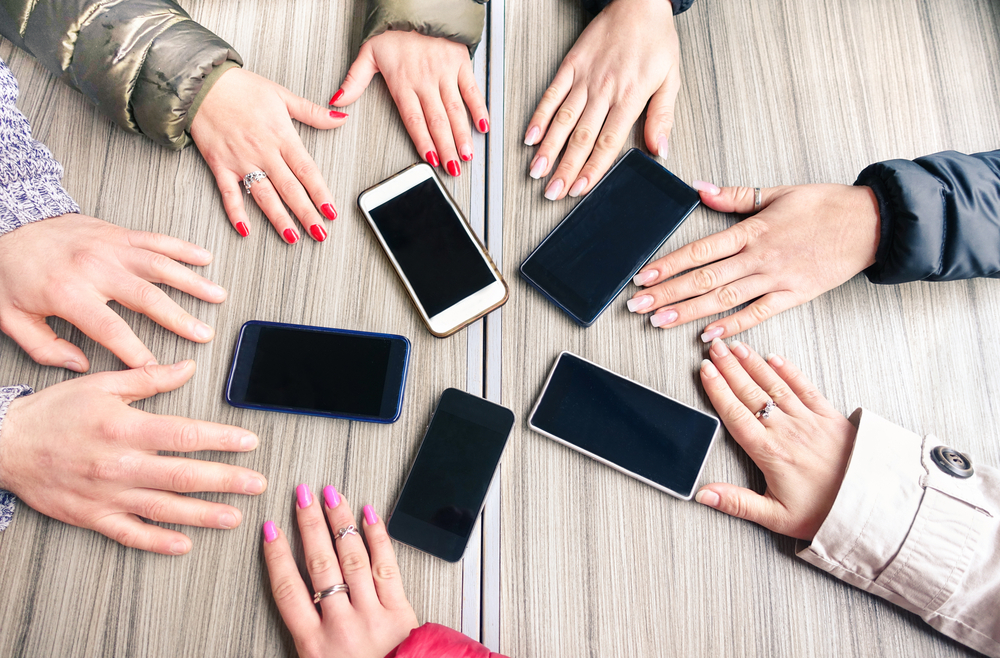 Group of friends having fun together with smartphones - Closeup of hands social networking with mobile cellphones - Technology and phone addiction concept on wood background - Focus on mobile phones
