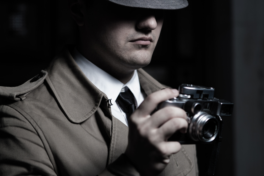 Young male spy agent wearing a hat, coat and a tie, holding rangefinder Leica film camera. Eyes hidden in the shadow.