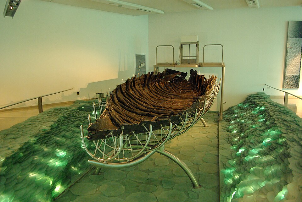 The 2,000-year-old fishing boat displayed in the Yigal Allon Museum with dramatic green lighting illuminating its preserved cedar planks and ancient construction.