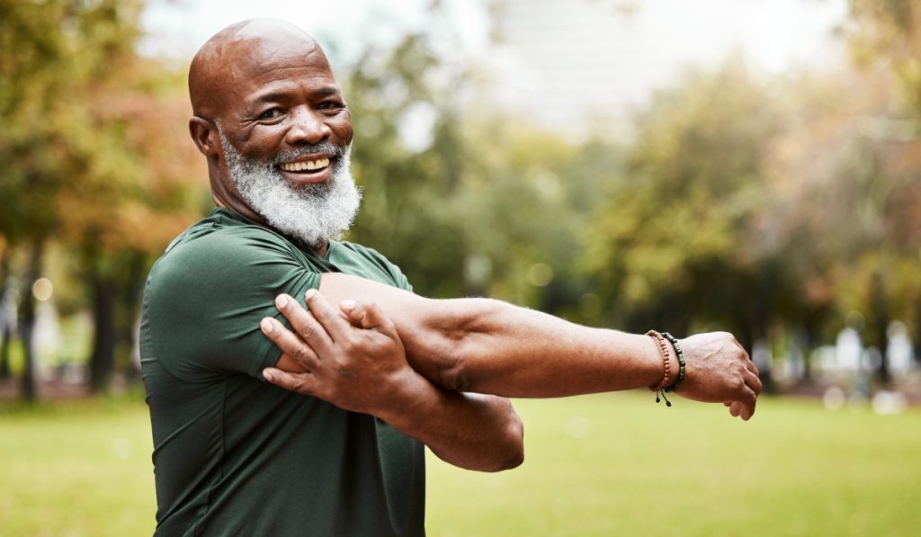 Senior fitness, exercise and black man stretching outdoor at park for energy, health and wellness in retirement. Portrait, face and smile of male in nature for workout, cardio and training in summer