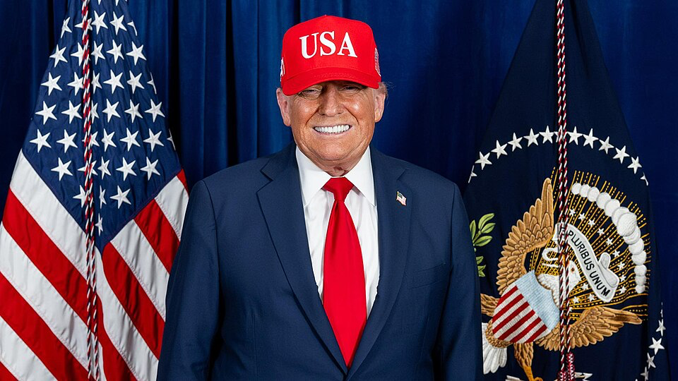 Donald Trump wearing red USA cap and dark suit on July 4th, 2025, standing in front of American flags and presidential seal
