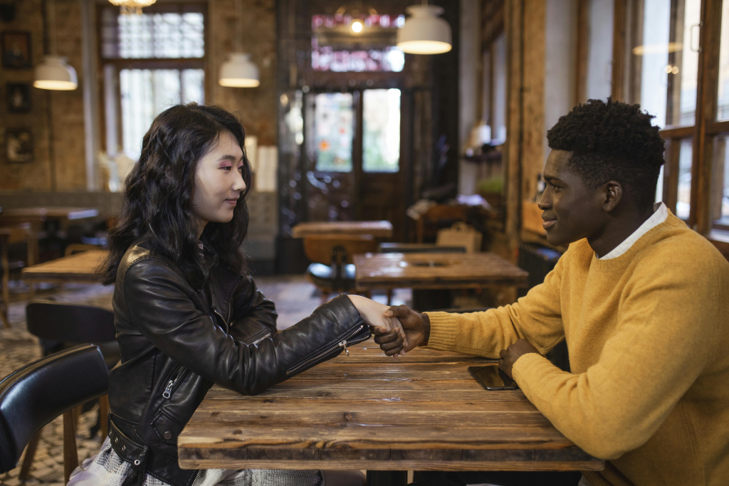 Two people hold hands across a wooden restaurant table, their connection depending on kissing compatibility.