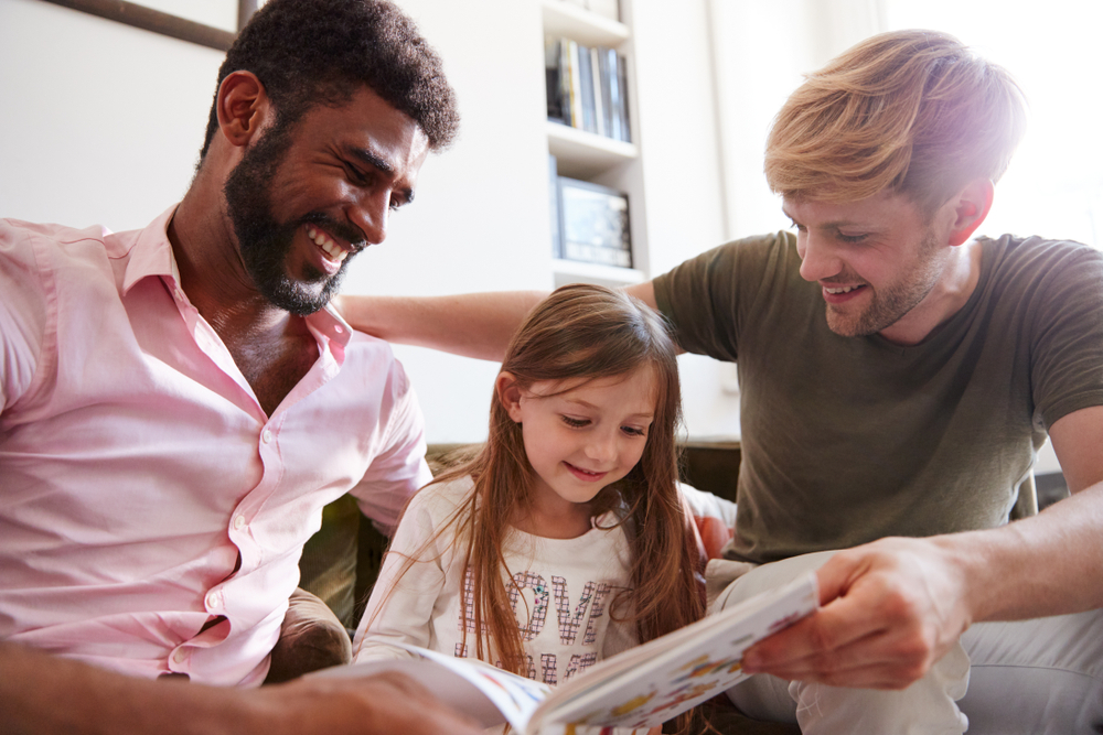 Same Sex Male Couple Reading Book With Daughter At Home Together Whilst Tickling Her
