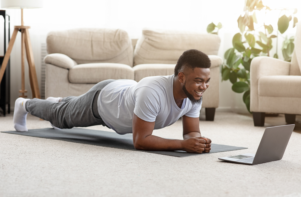 Training At Home. Sporty man doing yoga plank while watching online tutorial on laptop, exercising in living room, free space