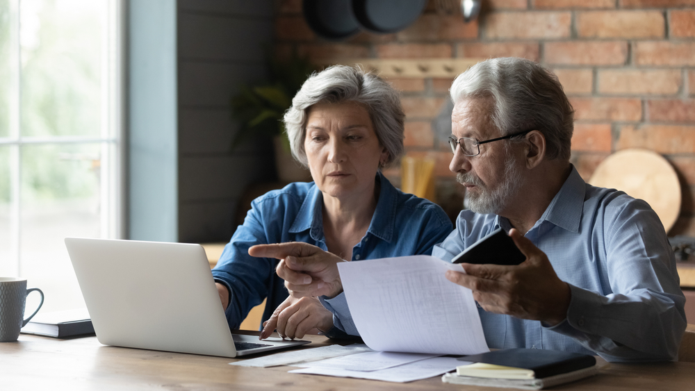 Focused senior husband and wife sit at table at home look at laptop screen pay bills taxes online. Concentrated mature man and woman couple make internet payment on computer, manage finances
