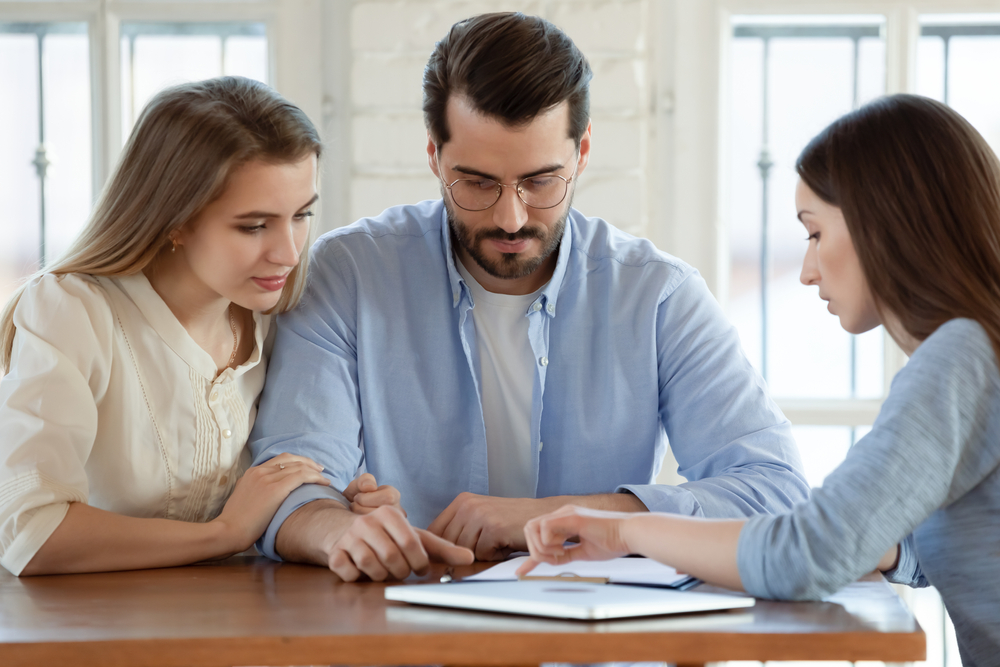 Focused young married couple discussing agreement with skilled real estate agent or broker. Professional financial advisor or saleswoman explaining contract details with interested clients at meeting.