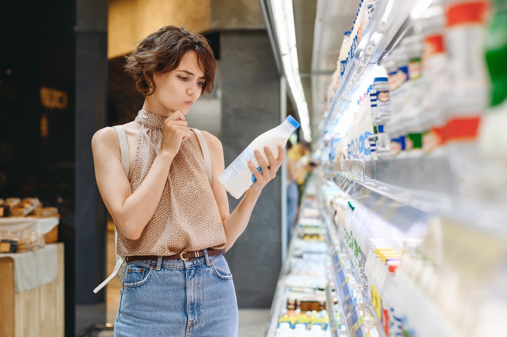 Young minded woman 20s in casual clothes backpack shopping at supermaket store buy choose dairy produce take milk read shelf life prop up chin inside hypermarket. Purchasing gastronomy food concept.
