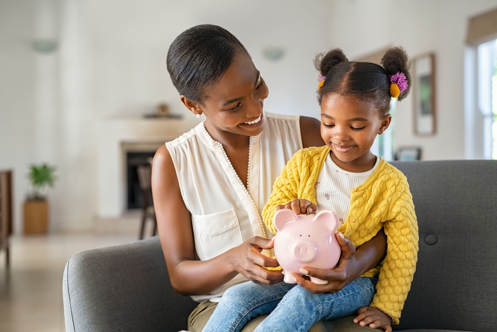 Smiling mature african american mother helping daughter sitting on lap putting money in piggy bank. Cute little black girl saving money by adding a coin in piggy bank with mother at home.