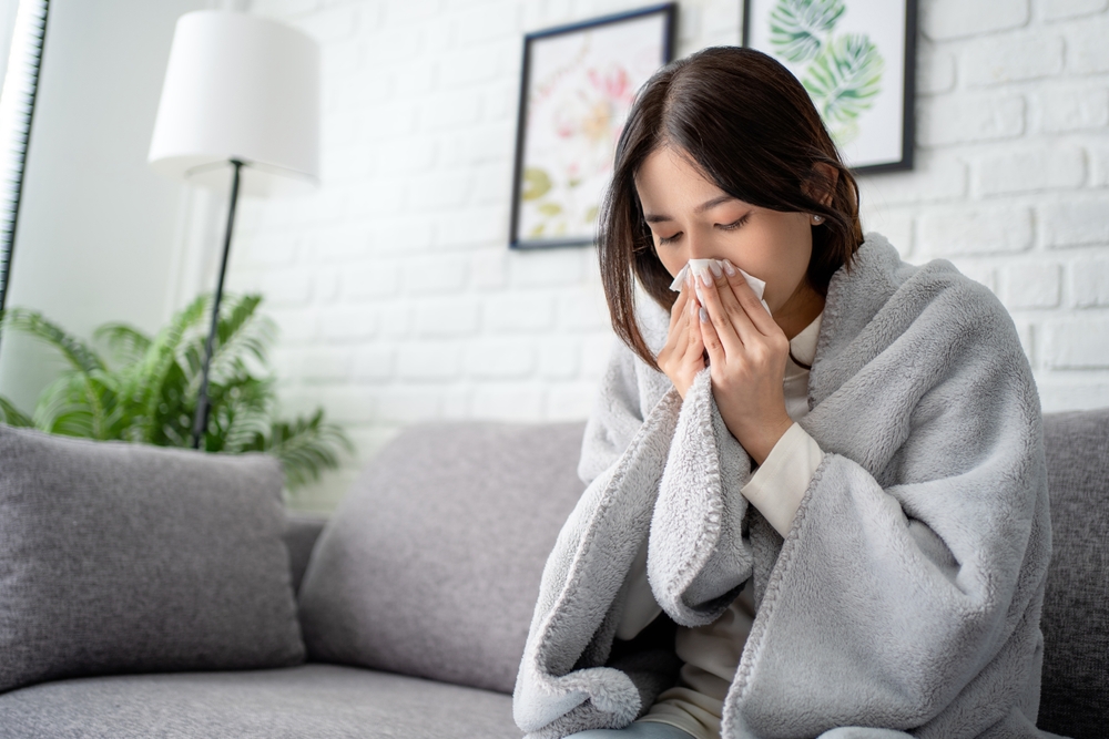 Sick young asian woman sitting under the blanket on sofa and sneeze with tissue paper at home.