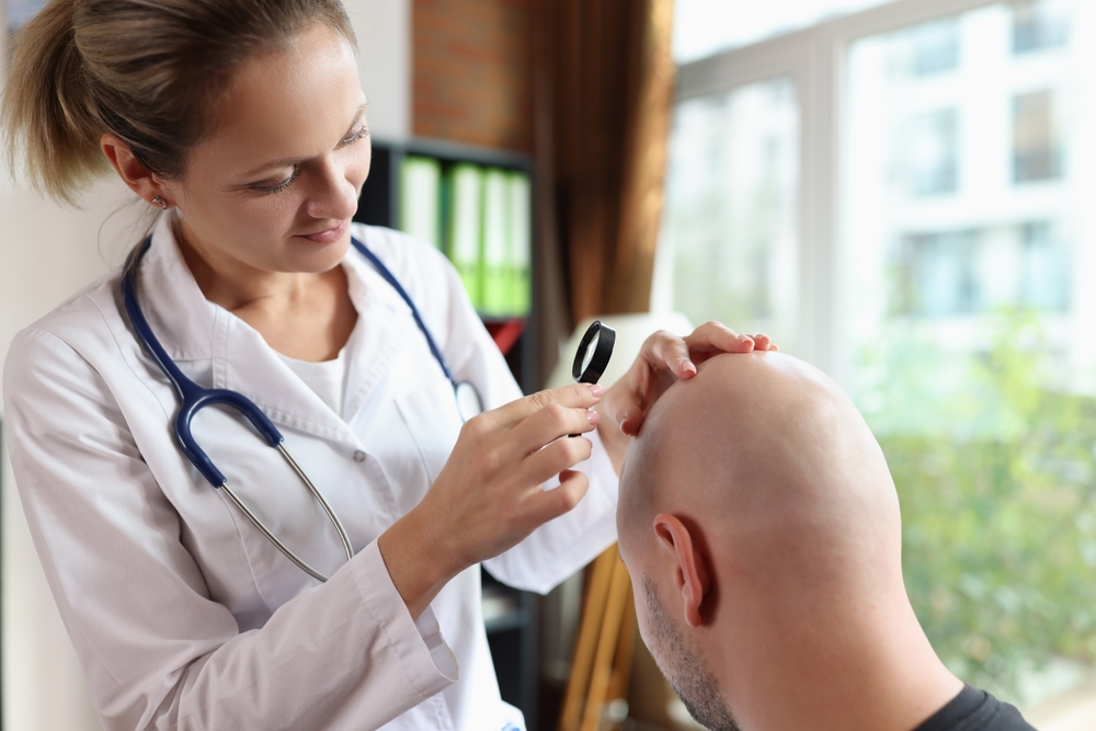 Female doctor with magnifying glass checks patient head skin in clinic office. Therapist examines man with alopecia symptoms. Skincare procedure