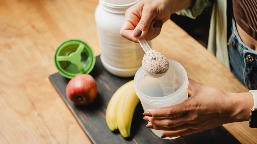 Woman in jeans and shirt with a measuring spoon in her hand puts portion of whey protein powder into a shaker on wooden table with white capsules, bananas and apple. Process of making protein drink.
