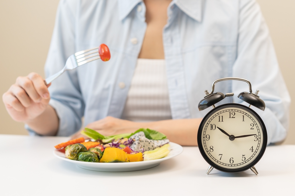 Intermittent fasting with clock, close up young woman, girl diet, waiting time to eat ketogenic low carb, green vegetable salad on plate. Eat food healthy first meal on brunch, lunch on table at home.
