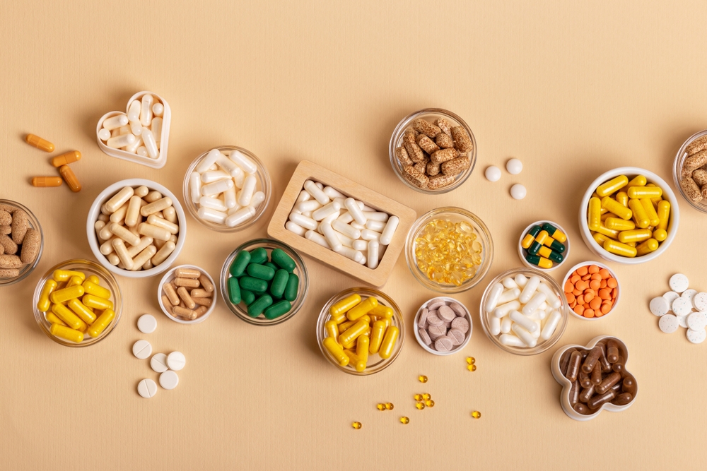 Many different colored vitamins, food supplements and minerals in glass jars, ceramic small plate and wooden box on a beige background top view. VItamin d, c, e, b, a, k, magnesium, zinc, probiotics.