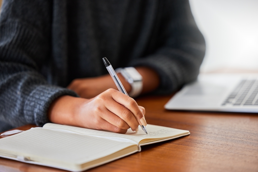 Notebook, writing and hand in home for entrepreneurship, small business and research with laptop. Female person, technology and journal in house for online research, remote work and meeting agenda