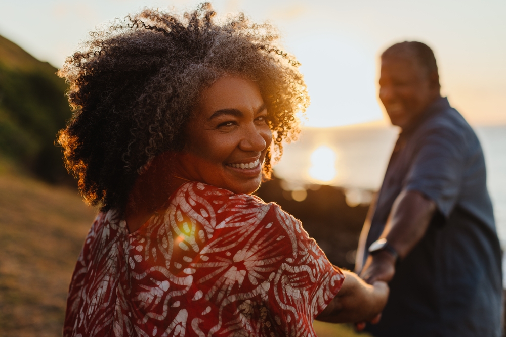 Senior woman embracing longevity and vitality while walking at sunset, holding her partner’s hand and smiling warmly.
