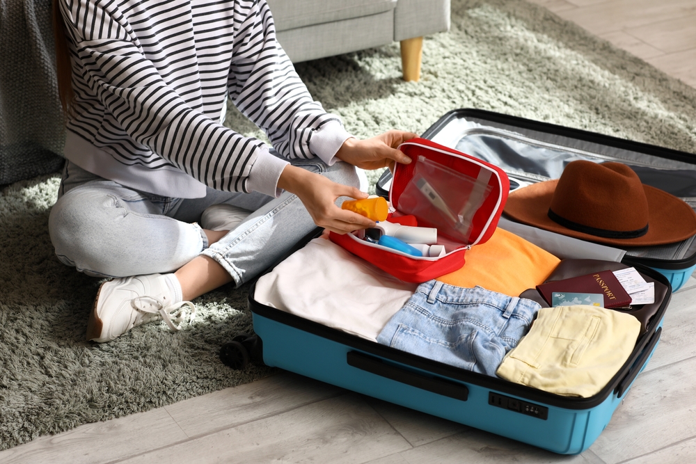 Young woman with suitcase packing first aid kit at home, closeup. Travel concept