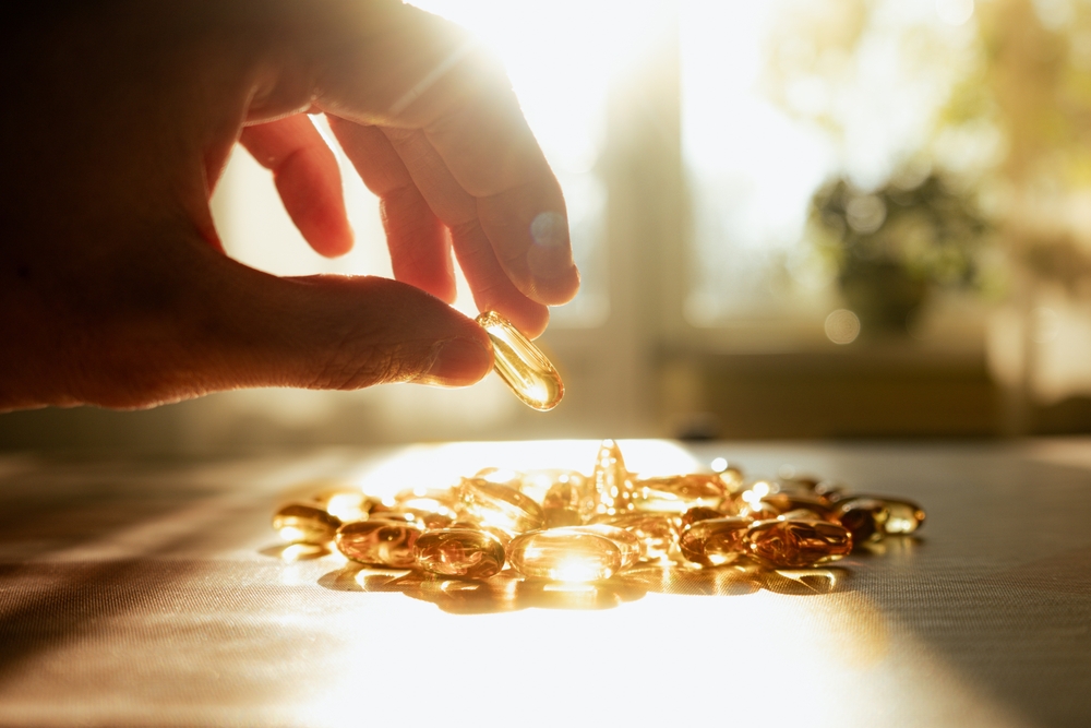 A man's hand reaching for yellow transparent vitamin D and Omega-3 fish oil capsules in the sunlight on a bright, sunny table.
