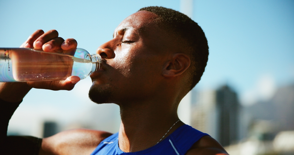 Fitness, black man and drinking water in city with fatigue, break and relax for exercise recovery. Workout, hydration bottle and thirsty athlete outdoor for wellness, body health or electrolytes
