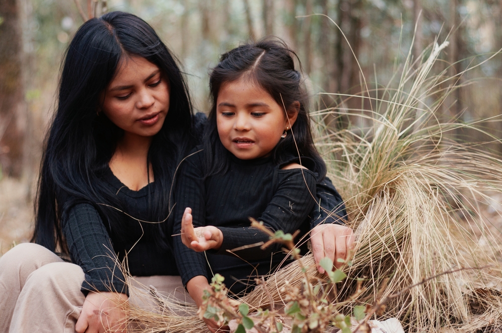 Indigenous mother and daughter are sitting together in nature, exploring the forest and discovering the local wildlife, creating a heartwarming moment of connection and learning