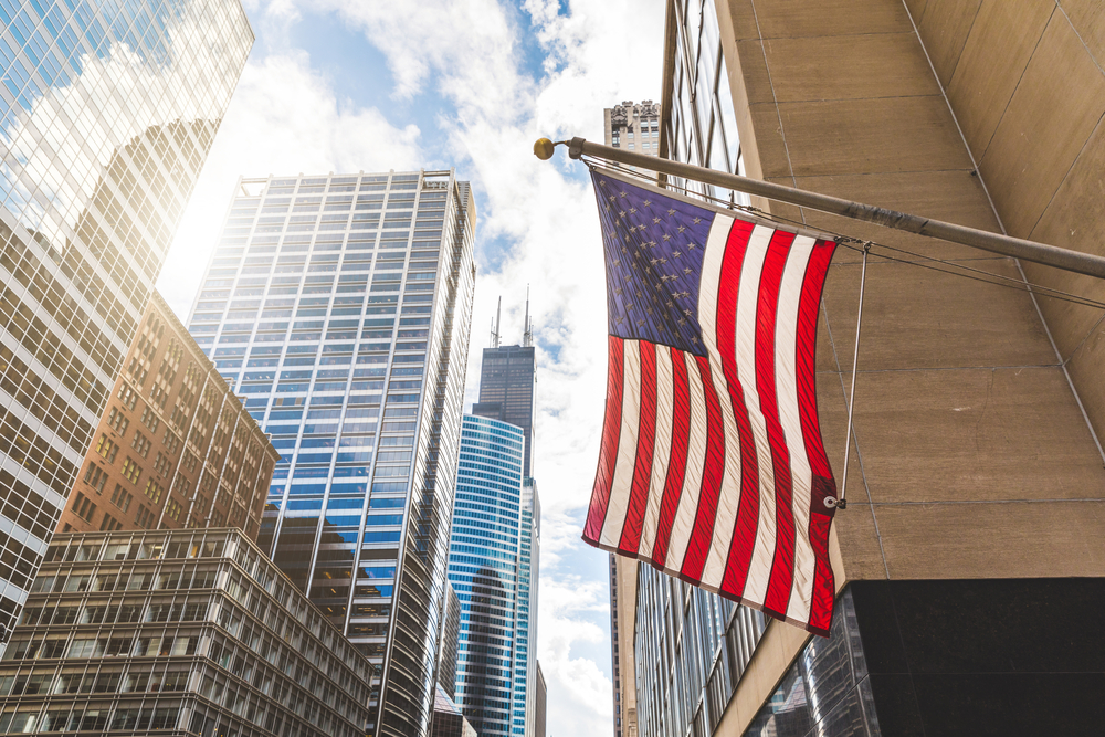 USA flag in Chicago with with skyscrapers on background. American flag waving in the city on a sunny day. Clouds reflections on buildings facade
