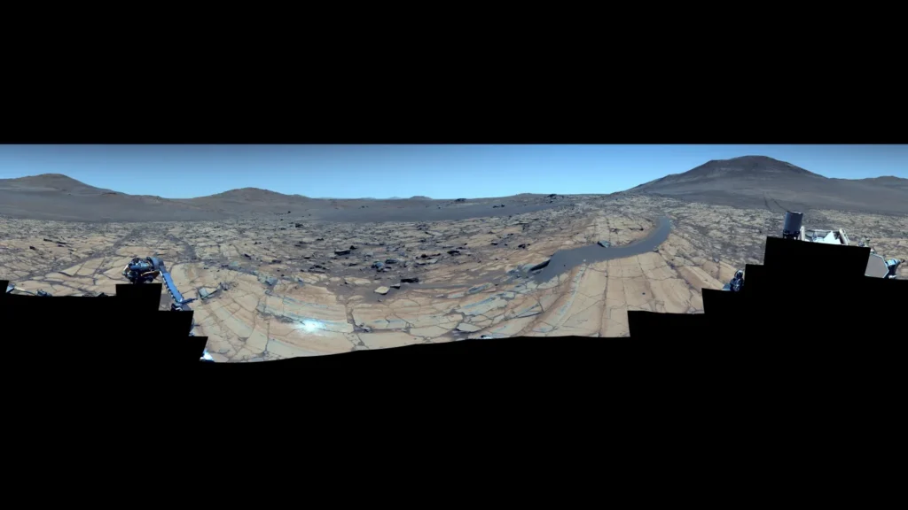 Wide panoramic view from Mars showing rocky terrain with scattered boulders under enhanced blue skies, with distant mountains on the horizon and rover tracks visible curving across the landscape.