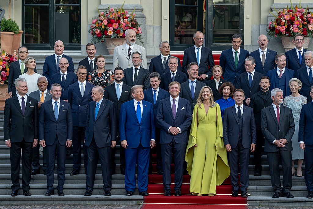 Trump stands with European leaders and officials on the steps of a formal government building during the NATO summit, with floral arrangements visible and participants arranged in multiple rows for the group photo.