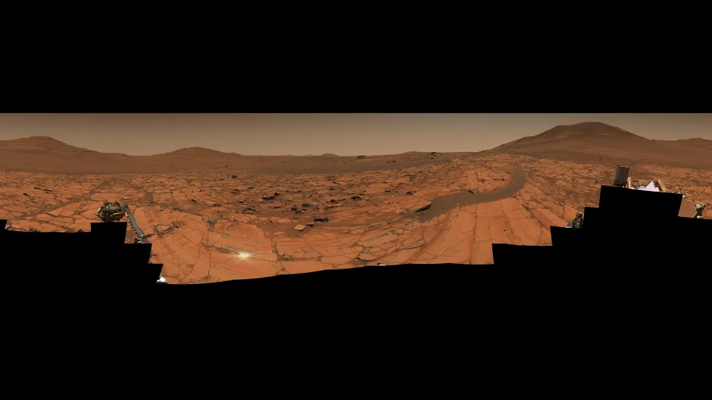 Mars panorama displaying the planet's natural rusty orange and amber tones across a boulder-strewn landscape, with rolling hills and distant peaks under the characteristic dusty atmosphere.