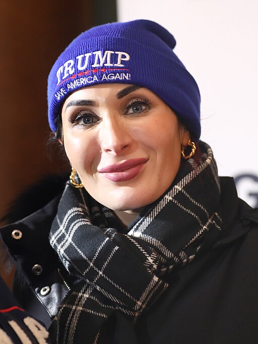 Laura Loomer wearing a blue "Trump Save America Again" campaign hat and plaid scarf at a political event in Des Moines, Iowa