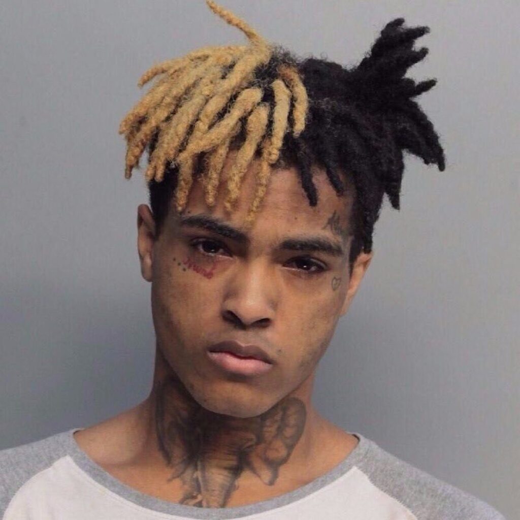 Booking photograph of XXXTentacion, showing the rapper with blonde and black dreadlocks, visible facial bruising around both eyes, neck tattoos, wearing a light gray shirt against a gray background.