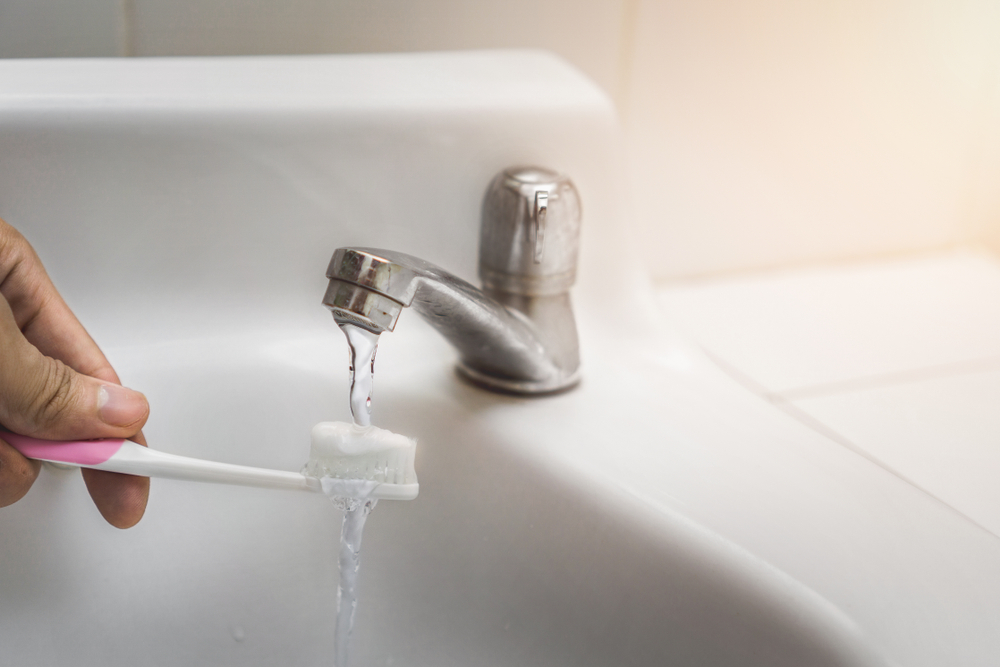 Close up image of a hand holding a pink toothbrush with Water being poured on toothpaste on top in bathroom sink.
