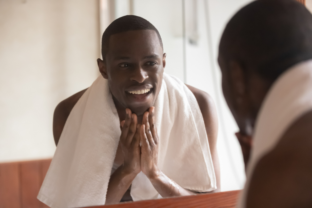 Black man towel on shoulders standing in bathroom look in mirror washes face touch skin after shaving or applying cream aftershave lotion feels satisfied, morning routine, personal hygiene, skincare