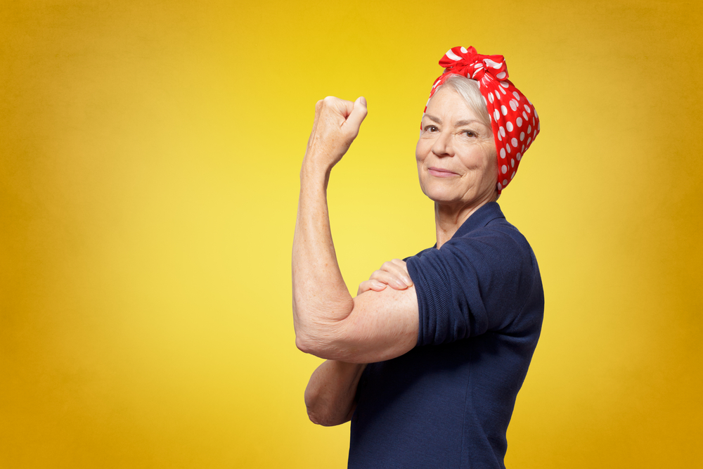 Rosie Riveter concept: proud senior woman with red headscarf, flexing her muscles, copy space, yellow background