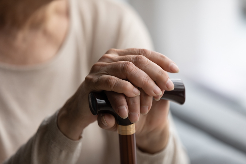 Close up focus on folded wrinkled female hands on wooden cane. Cropped thoughtful elderly senior grandmother having walking disability, using stick indoors, old retired people lifestyle concept.