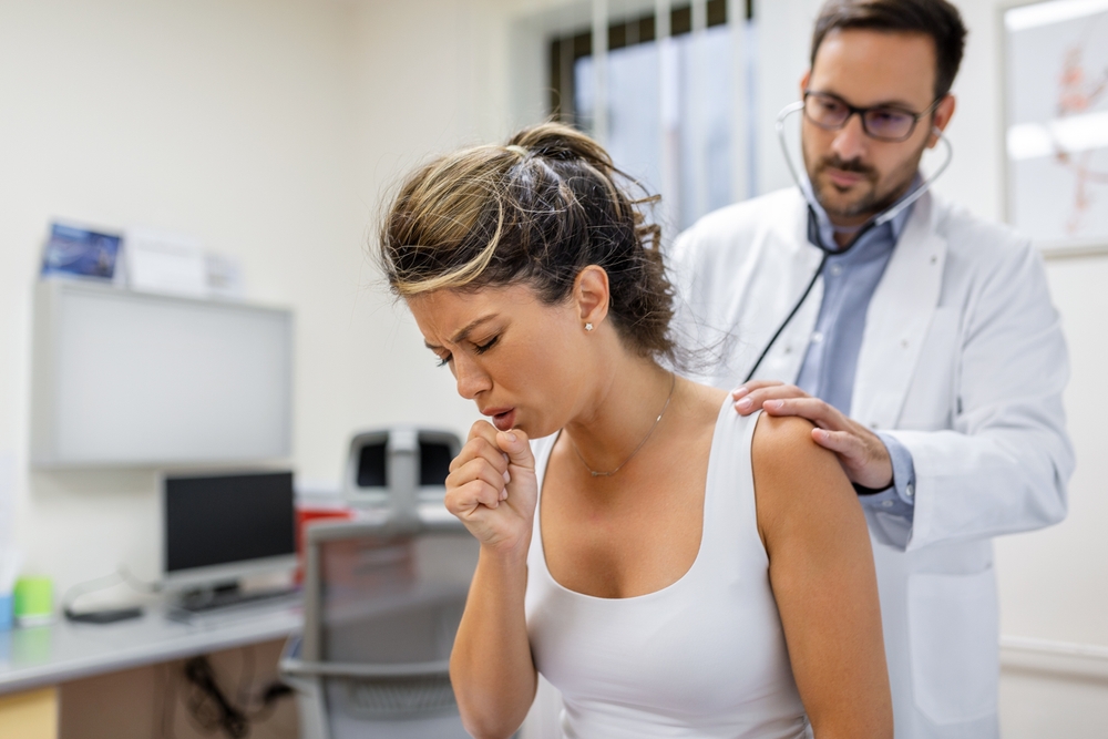 Young female patient in the clinic suffered from pneumonia, she is coughing the doctor listens to the wheezing in the lungs with a stethoscope.