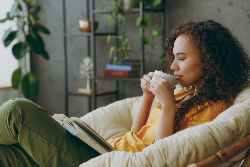 Sideways young woman of African American ethnicity wear t-shirt drink coffee read book close eyes sits in armchair stay at home flat rest relax spend free spare time in living room indoor grey wall
