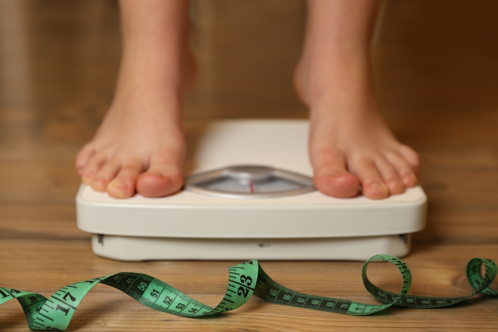 Overweight girl using scales near measuring tape on wooden floor, selective focus

