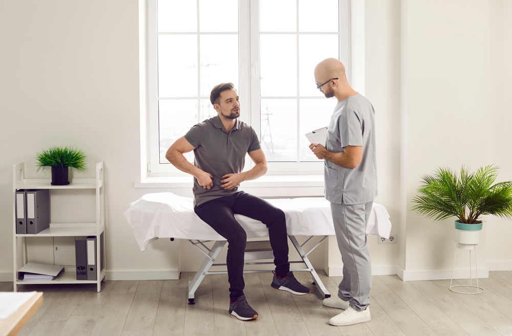 Young man sitting on the couch in the doctor's office and pointing to his stomach to the doctor during medical examination in clinic. Physician listening to the patient's complaints.
