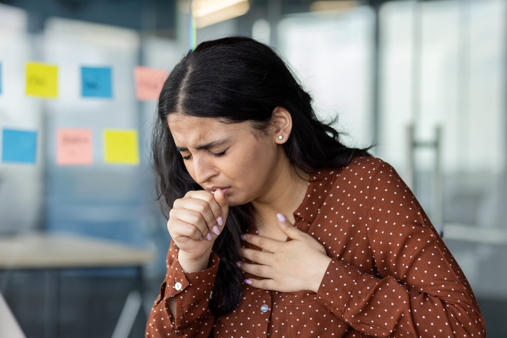 Sick woman close up coughing at workplace, businesswoman holding hand to chest, having cold. Office worker inside office at workplace.
