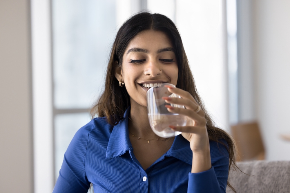 Attractive Indian woman holding glass of still or mineral water seated on sofa at home, making sip, enjoy refreshment, prevents body and skin dehydration, flush out toxins, keeping healthy life habit