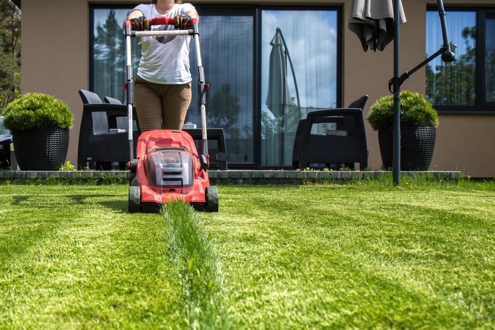 A woman mowing a lawn with a red lawn mower. The lawn mower is red with black elements.