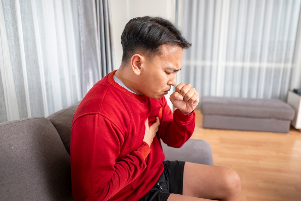 Indonesian southeast asian man sitting on the couch at home and coughing. Suffers from an attack of asthma or allergies. Healthcare concept