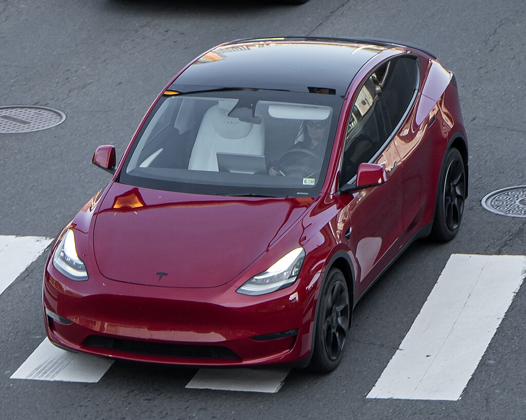 Aerial view of a red Tesla Model Y parked on a street with white crosswalk markings visible on the pavement.