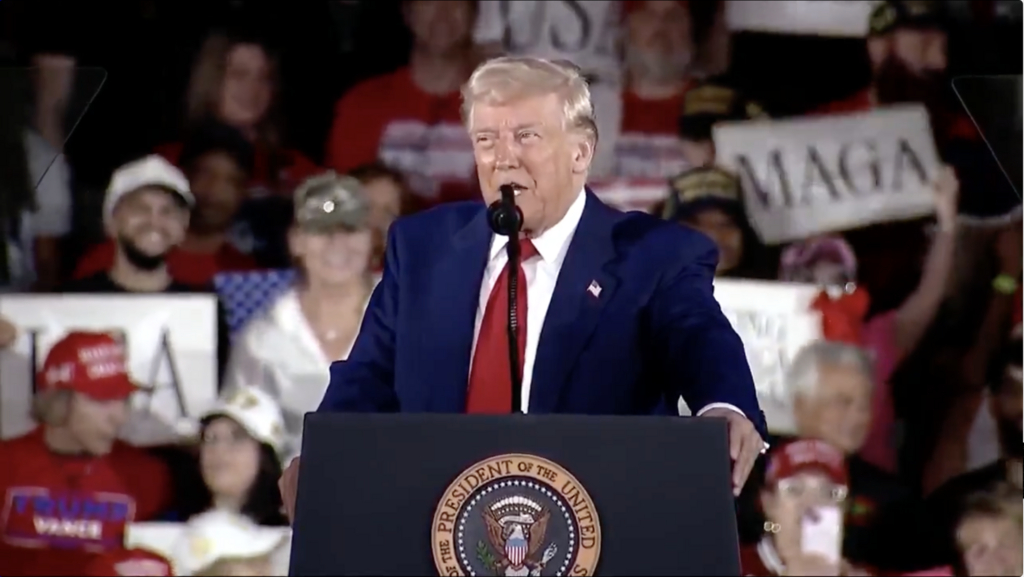Donald Trump speaks at a podium with the presidential seal, wearing a dark suit and red tie. Supporters behind him hold MAGA signs.
