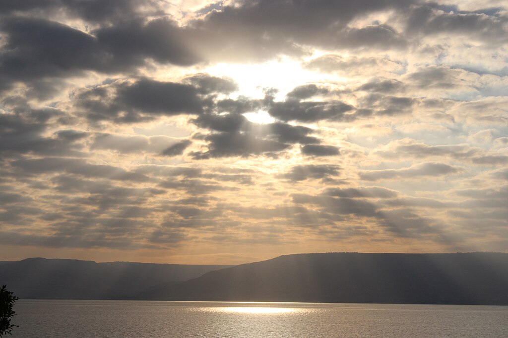 Dramatic view of the Sea of Galilee at sunset with sunbeams breaking through storm clouds over the water, mountains visible along the distant shoreline.